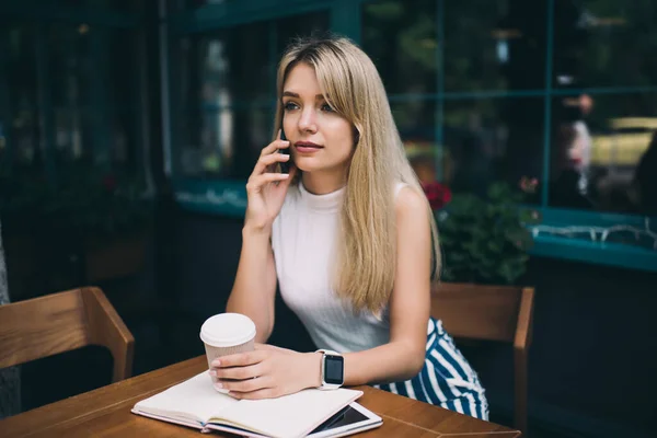 年轻的金发女人穿着无袖的高领毛衣 坐在咖啡店外面的桌子旁 一边打电话 一边一边一边手拿着一杯咖啡 — 图库照片