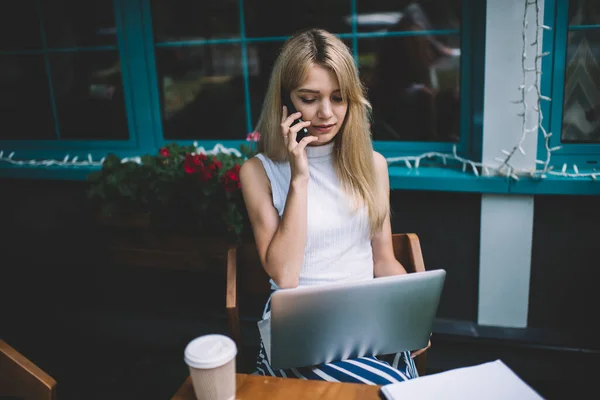 ブロンド深刻な笑顔スマート女性でノートパソコンでテーブルに座っている携帯電話で話している袖なしシャツで街のカフェテリア — ストック写真