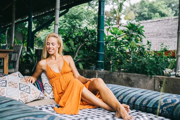 オレンジ色の夏のドレスを着た幸せな魅力的な女性は オープンエアのカフェで枕の上に横たわって笑顔とバリ島の休暇中に見下ろして — ストック写真