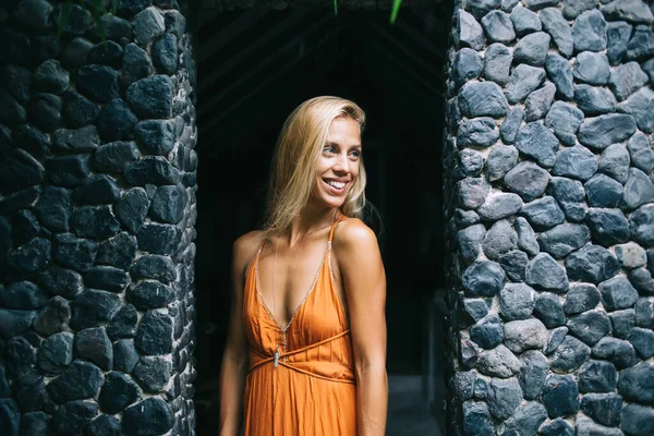 かなり日焼けしたブロンド女性とともに魅力的な笑顔身に着けています明らかにオレンジドレスとストラップと深いDecollet立っているダークグレーの石の壁の横に — ストック写真
