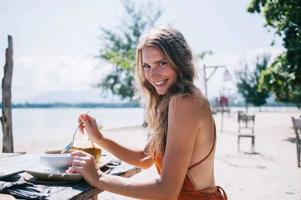 オレンジ色のドレスを着た魅力的な大人のブロンドの女性の側面図カメラを見て笑顔素朴な木に座っている間 バリのリゾートで海沿いのおいしい料理と新鮮なココナッツドリンクとテーブルを提供晴れた日の間に — ストック写真