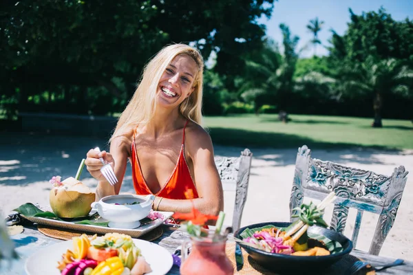 Pretty adult woman in orange clothes looking at camera and laughing while sitting at served table with plates of colorful fresh exotic fruit mix and beverages and holding fork on background of blurred green garden at Bali resort