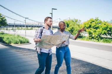 Coşkulu çoklu etnik arkadaşlar haritayla bölgeyi keşfediyor ve yeşil ağaçlar ve köprü inşaatının arkasındaki boş yolda yürüyorlar. 