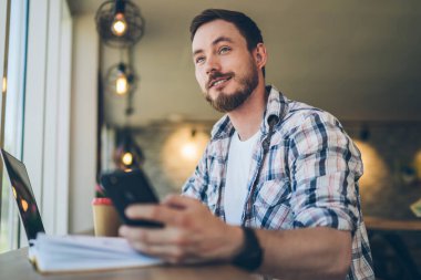 İş yerinde cep telefonuyla sörf yaparken kareli tişörtlü, güler yüzlü, düşünceli, düşünceli bir erkeğin bakışlarının altından.