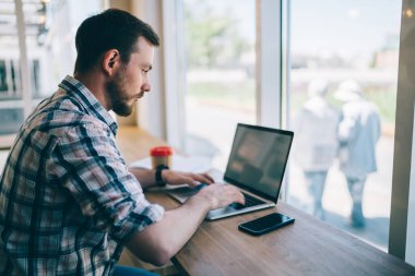 Gömleği kollu, odaklanmış sakallı bir adam bir fincan kahve alıp kafeteryada bilgisayarla uzaktan kumandayla çalışıyor.