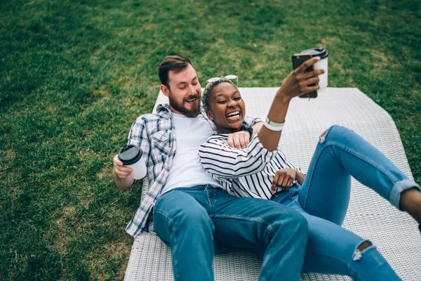 松了口气的多民族夫妇躺在垫子上 手里拿着纸杯 热饮男朋友从上面拥抱着兴奋的非洲裔美国女朋友 — 图库照片
