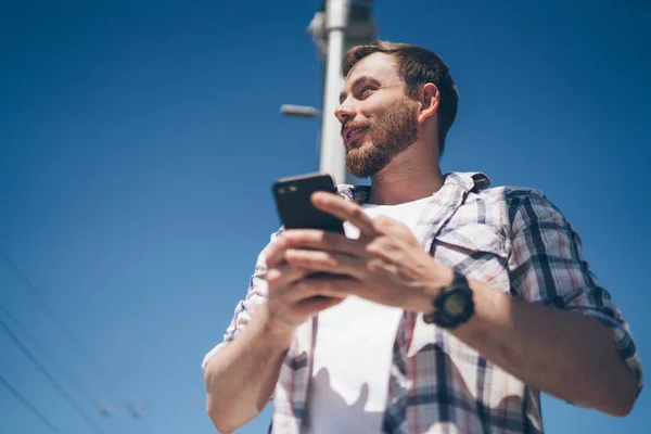 From below of positive young bearded male in casual clothes smiling and looking away while standing on street with smartphone in hands on summer day with blue sky on background