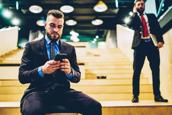 穿着正装的文雅而严肃的男人与朋友坐在现代建筑的楼梯上 用手机聊天来放松自己 — 图库照片