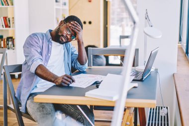 Yorgun sakallı siyah adam alnına dokunuyor ve masa başında oturmuş, ofis planları üzerinde çalışırken kameraya bakıyor.