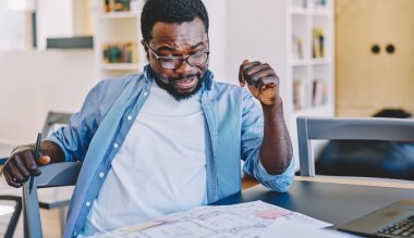Gündelik kıyafet ve gözlüklü etnik adam, masa başında otururken ve modern işyerinde proje üzerinde çalışırken taslak gözden geçiriyor.