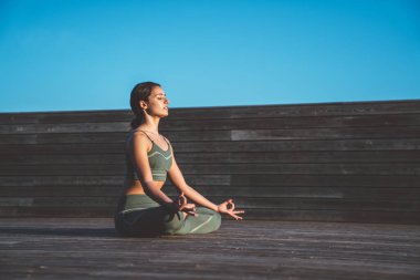 Yeşil spor elbiseli, sakin etnik zayıf bir kadının Lotus duruşu içinde el ele tutuşup ahşap terasta gözlerini kapamasının yan görüntüsü.