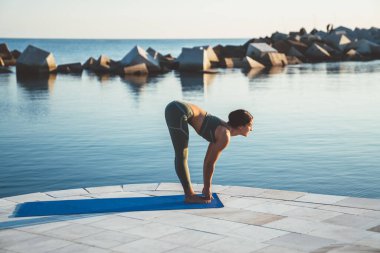 Yeşil spor giyim giymiş esnek kadın sabah deniz suyunun yanında mavi hasırda yarı kaldırma pozisyonunda yoga yapıyor.