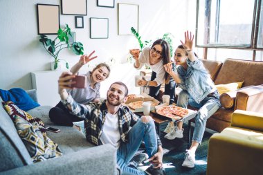 Neşeli bir grup arkadaş canlısı, gülümseyen ve cep telefonuyla selfie çeken bir grup insan masada, koltukta ve yerde bacak bacak bacak üstüne atarak pizza yiyorlar. 