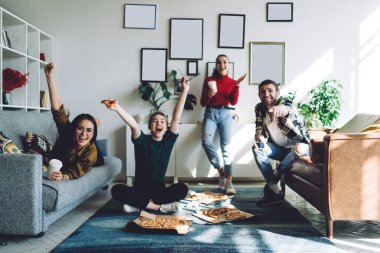 Heyecanlı genç grup, televizyonda maç izlerken ve rahat bir dairede pizza yerken farklı jestlerle başarıyı kutluyor.