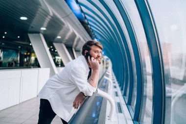 Hafif geometrik koridorda çitlere yaslanmış, cep telefonuyla konuşan hoş sakallı adamın yan görüntüsü.