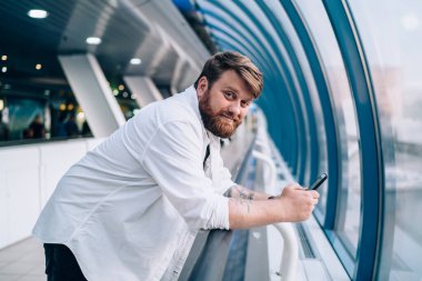 Modern cep telefonu cihazını halka açık bir yerde dinlenirken kameraya bakan, cep telefonuyla sohbet eden, cep telefonlu yakışıklı hippi portresi.