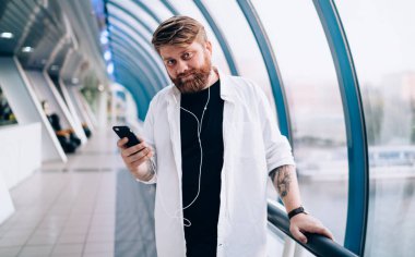 Elektronik kulaklıklı yakışıklı hippi adamın yarım boy portresi. Kapalı mekanda kameraya bakıyor ve öğrenmek için ses kitabı dinliyor. Cep telefonlu, boş zamanlarında poz veren sakallı bir adam.