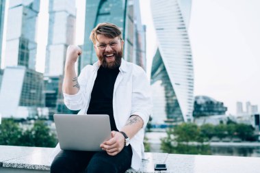Saati ve dizüstü bilgisayarı olan, günlük kıyafetleriyle mutlu bir erkek iş bölgesinde çitlerin üzerinde başarılı bir projeyi kutluyor.