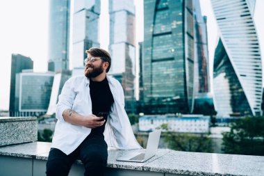 Gözlüklü, beyaz tişörtlü, cep telefonuyla mesajlaşan mutlu sakallı bir adam. Gökdelenleri olan bir barajın yanında duruyor. 