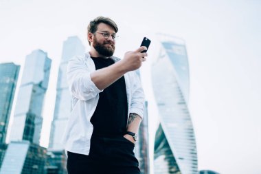 Düşük açılı mutlu, dolgun gözlüklü, sıradan kıyafetler giymiş, selfie yapan, arka planda modern ayna gökdelenlerinin karşısında duran bir adam. 