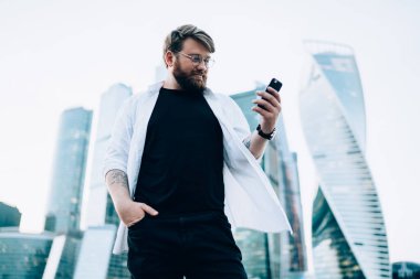 Gözlüklü, beyaz tişörtlü ve siyah pantolonlu gülümseyen bir erkek modern şehirde cep telefonunda geziniyor. 