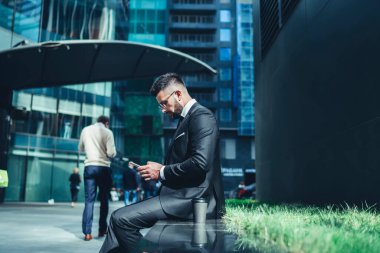 Ekrana konsantre olan ve şehir merkezindeki bankta otururken akıllı telefon kullanan düşünceli, kendini işine adamış bir çalışanın bakış açısından.