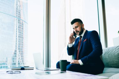Takım elbiseli sakallı bir erkek, telefona cevap veriyor ve güncel ofise gitmek için laptop ve içeceklerle masanın yanındaki koltukta otururken düşünüyor.