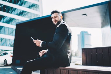 Siyah takım elbiseli, cep telefonuyla gezen şık bir erkeğin yan görüntüsü. Şehirde, güneş ışığında ahşap bir podyumda oturuyor.