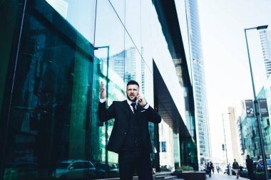 Koyu renk takım elbiseli deli adam şehir merkezindeki çağdaş binanın dışında akıllı telefondan konuşurken el kol hareketi yapıyor ve başka tarafa bakıyor.