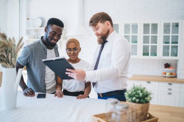 Profesyonel sakallı, resmi kıyafetli şişman adam modern güneşli dairede mutfak masasının yanında dururken ilgilenen etnik çifte evrak gösteriyor.