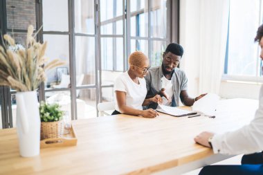 Düşünceli Afrikalı Amerikalı çift, emlakçının önünde masada oturmuş, emlak sözleşmelerini dikkatle okuyor ve tartışıyorlar.