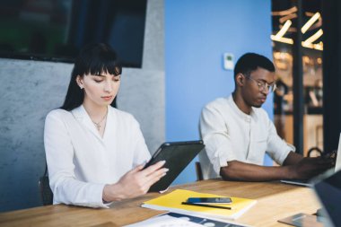 Rapor çalışırken ve modern iş merkezindeki masada otururken beyaz tişörtlü, tablet ve laptop kullanan çok ırklı, hüzünlü ortaklar.