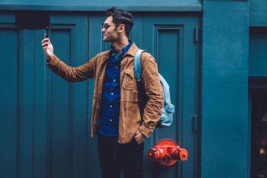 Sırt çantalı, modaya uygun giysileri ve güneş gözlükleriyle gülümseyen genç adam New York caddesinin mavi binasında dururken selfie çekiyor.