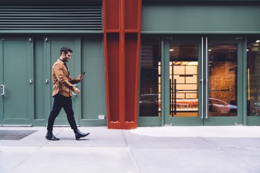 New York caddesindeki binaya karşı şık kıyafetler içinde yürüyen ve akıllı telefondan yazan gülümseyen genç bir erkek.
