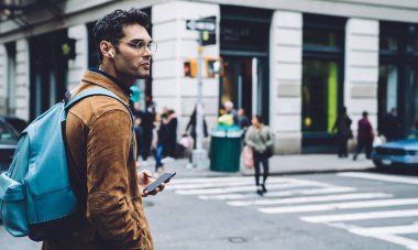 Ceketli, kulaklıklı, siyah saçlı, mavi sırt çantalı, New York caddesinde yürüyen yakışıklı bir adam.