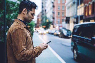 Ceketli ve gözlüklü, akıllı telefon kullanan, güpegündüz trafik şeridinin yanında duran kendinden emin, yakışıklı, esmer bir adam.
