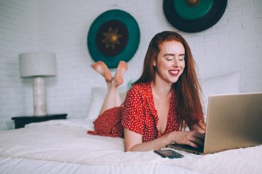 Romantik, mutlu, kırmızı saçlı, parlak noktalı elbiseli bir kadın evde akıllı telefon ve dizüstü bilgisayar kullanırken rahat bir yatakta uzanıyor.