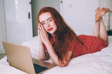Düşünceli kızıl saçlı, kırmızı noktalı elbiseli, minimalist ışık konutlarında dizüstü bilgisayar kullanırken rahat bir yatakta poz veren ve kameraya bakan düşünceli bir kadın.
