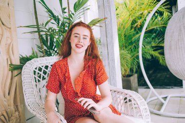 Kırmızı benekli elbiseli, güzel, kızıl saçlı kadın bahçe sandalyesinde dinleniyor cep telefonunu güvertede tutuyor ve kameraya bakıyor.