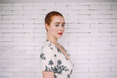 Beyaz tuğla duvarın önünde dikilip kameraya bakan yaz elbiseli, güler yüzlü kızıl saçlı kadının yan görüntüsü.