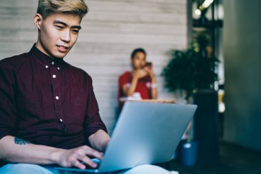Odaklanmış Asyalı erkek, sarı saçlı, eflatun tişörtlü dizüstü bilgisayarda dizlerinin üzerinde yaratıcı çalışma alanında oturan ve arka planda meslektaşı olan.