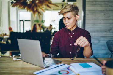 Odaklanmış ciddi Asyalı erkek, sarı saçlı, günlük eflatun gömlekli, gözlük takan, yaratıcı iş yerinde dizüstü bilgisayar kullanan, arka planda meslektaşları olan.