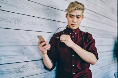 Konsantre Asyalı erkek, sarı saçlı, günlük kestane rengi gömlekli, gözlük takan, ahşap duvara yaslanmış cep telefonunu izleyen.