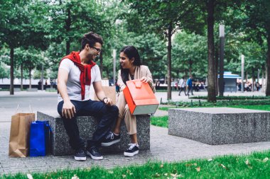 Bir çift kağıt torbaların yanında taş bankta otururken, bir kadın dışarıda parkta alışveriş yaptıktan sonra bulanık arka planda alışverişlerini gösteriyor.