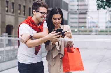 Moda tebessümlü, modern, çok ırklı genç adam ve kadın alışveriş çantasıyla akıllı telefonlara göz gezdirirken gün ışığında şehir kaldırımında duruyorlar.