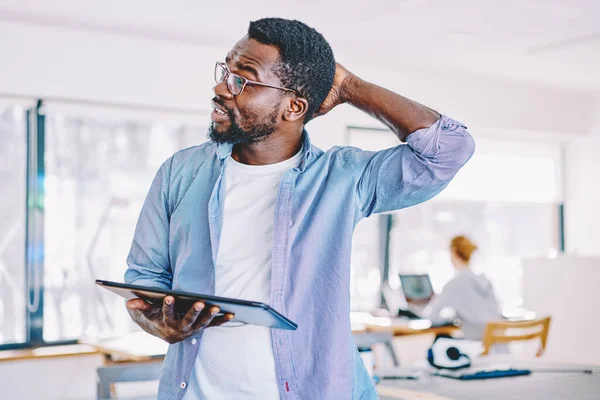 Puzzled black man in casual outfit and with tablet scratching head and thinking over problem while working on project in modern office