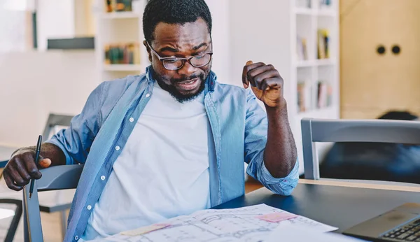 Gündelik kıyafet ve gözlüklü etnik adam, masa başında otururken ve modern işyerinde proje üzerinde çalışırken taslak gözden geçiriyor.