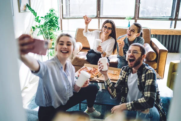 Group of delighted young people with pizza and beverages to go smiling and taking selfie while sitting near table during gathering at home