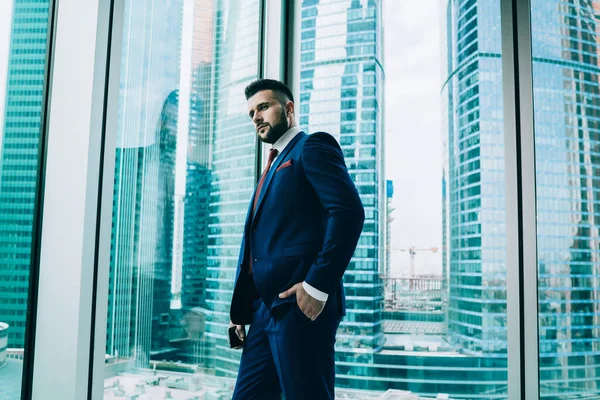 From below pensive well dressed executive man with smartphone looking out window and pondering after hard phone conversation in modern office of high rise building