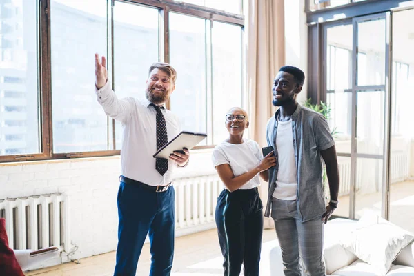 Positive bearded chubby man in formal outfit with clipboard showing living room to smiling amused ethnic couple while standing at modern sunny apartment